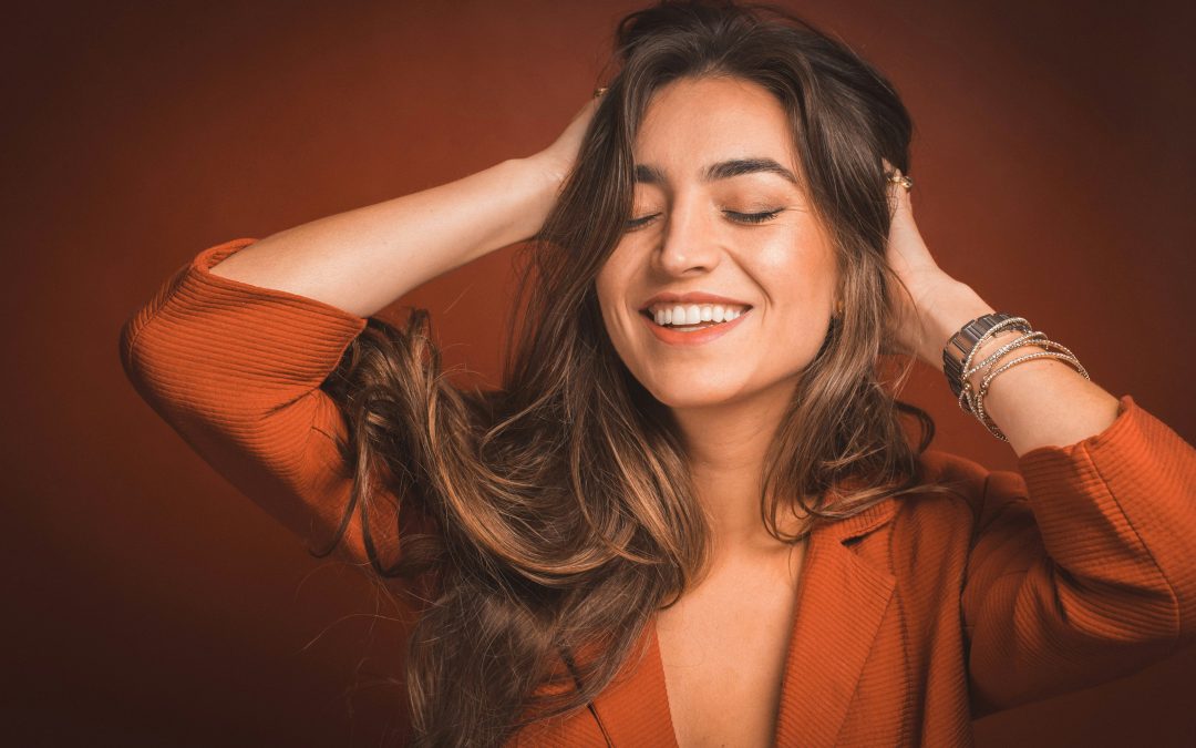 Femme aux cheveux longs et brillants, souriante, portant une veste orange, illustrant les bienfaits des masques capillaires
